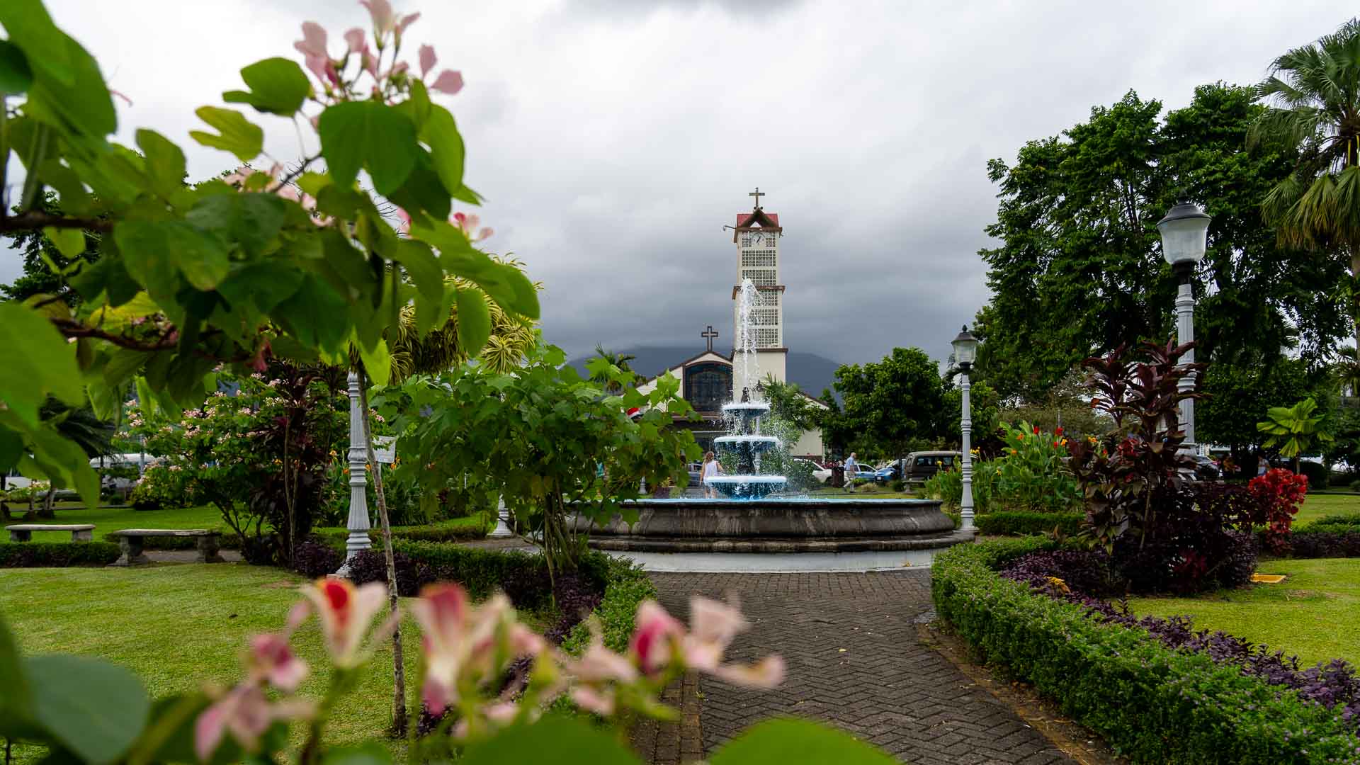 Water shoots out of a fountain in front of a church. The church is located in a flowery garden.
