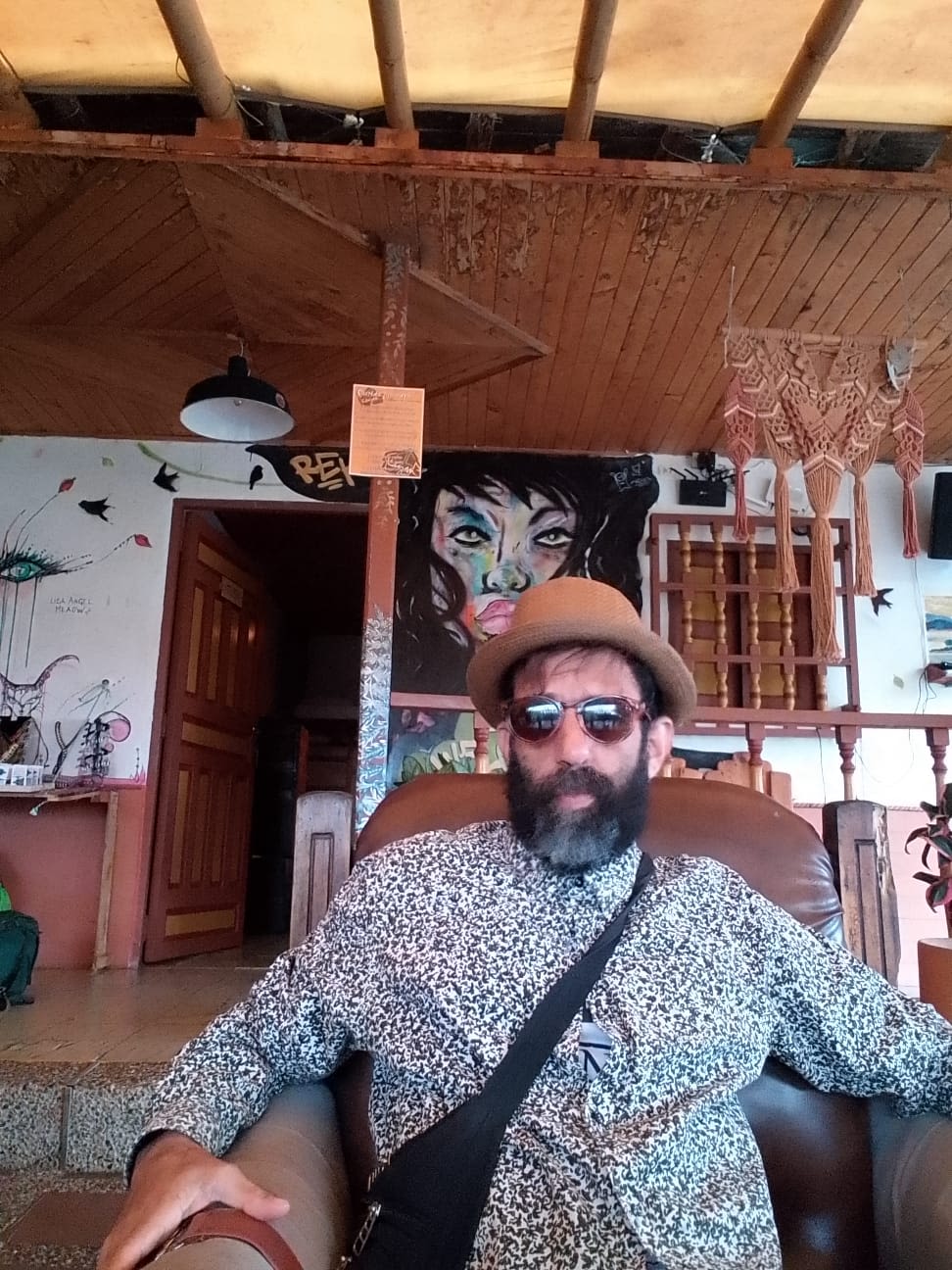 A man sat in a chair looks at the camera. He is wearing a hat and sunglasses.